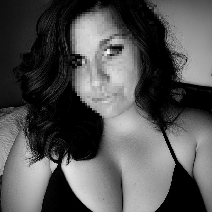 Black and white image of a curvy woman with dark, wavy hair cascading over her shoulders, wearing a black bra. Her face is pixelated, maintaining the allure of anonymity. The softly lit bedroom setting enhances the intimate yet commanding presence of this